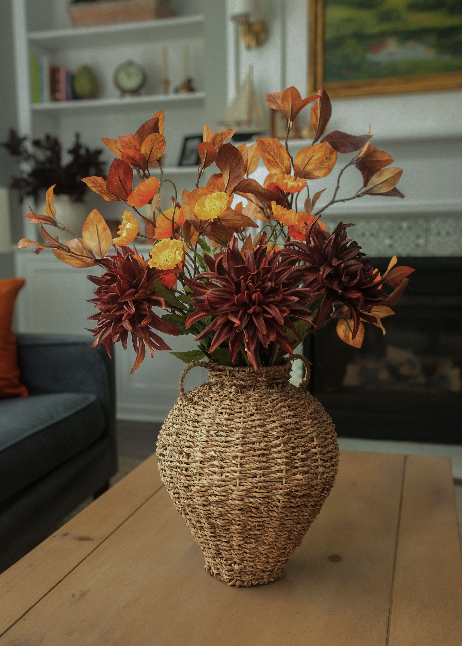 An artificial autumn floral arrangement in a woven straw vase, displayed on a wooden table in a living room setting.