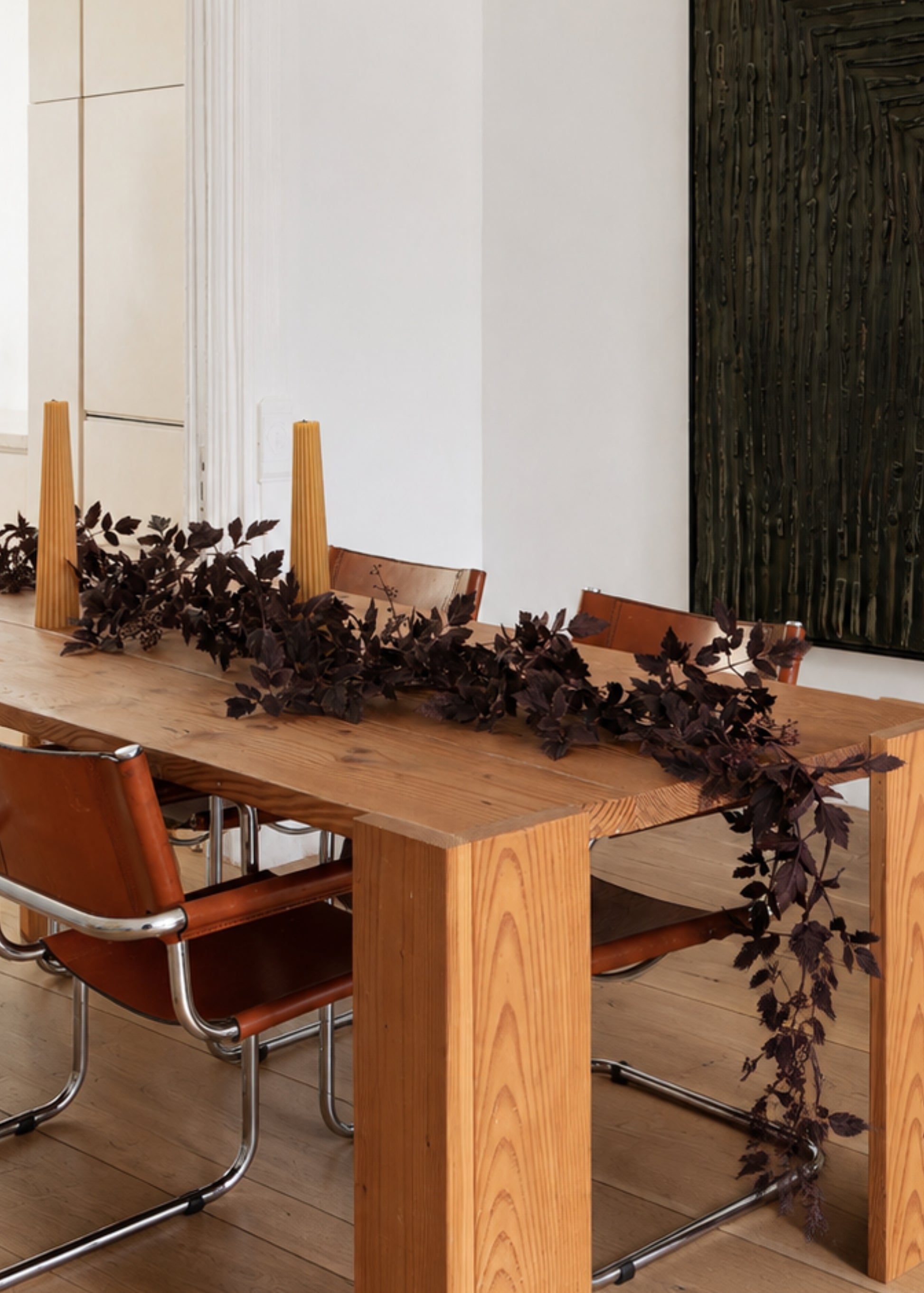 A rustic wooden dining table with a dark leaf garland and tall taper candles, surrounded by modern brown leather chairs in a bright interior.
