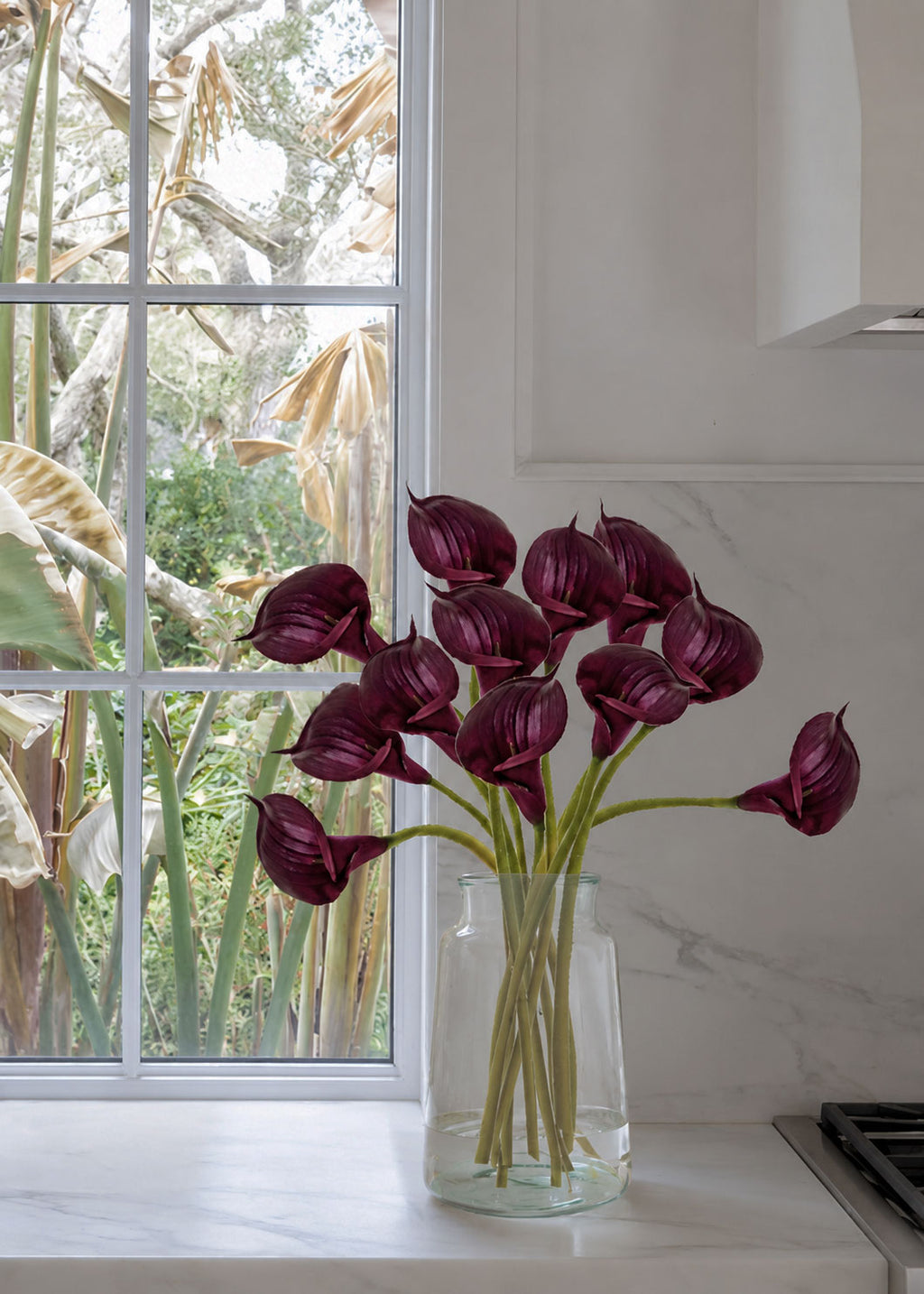 A bouquet of deep plum calla lilies in a clear glass vase, set on a white marble kitchen counter next to a window 