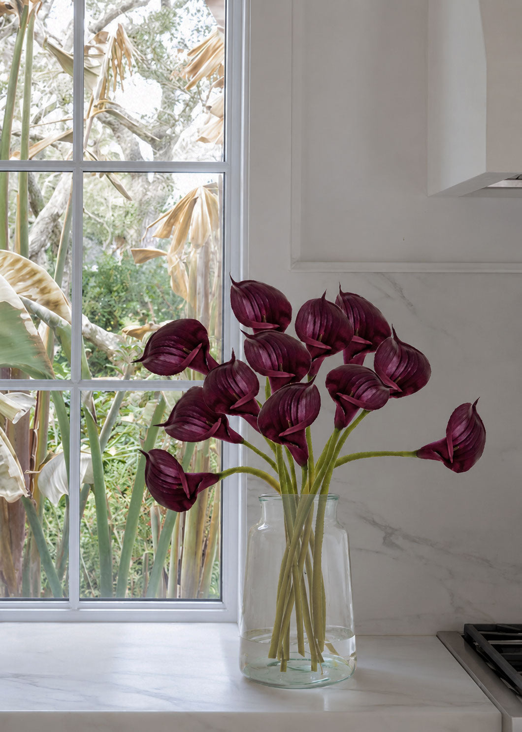 A bouquet of deep plum calla lilies in a clear glass vase, set on a white marble kitchen counter next to a window 