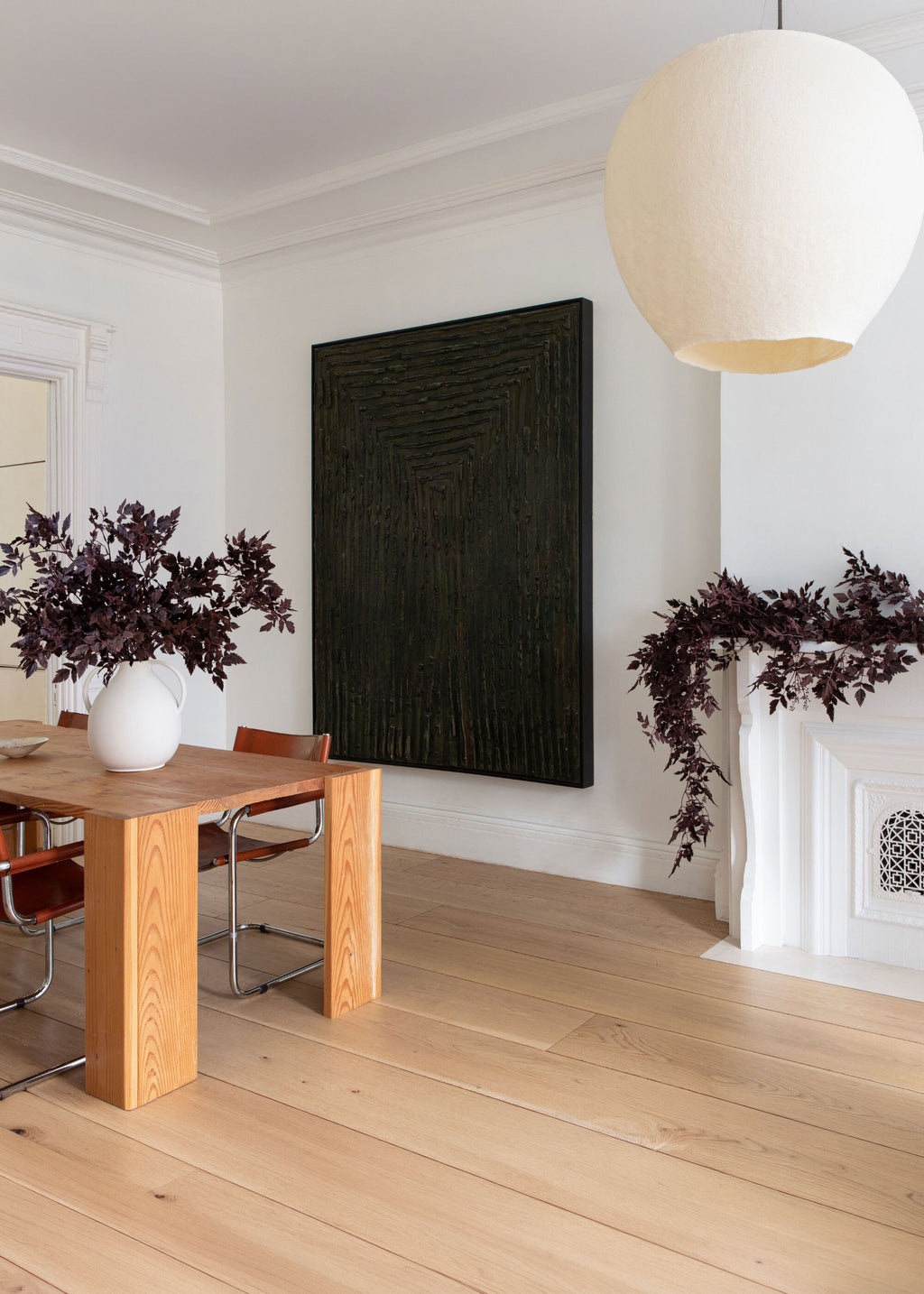 A large, vertically oriented black textured wall art piece hangs on a white wall in a modern dining room setting with light wood floors, a wooden dining table, chairs, a white pendant light, and faux cimicifuga leaf garlands and arrangement.