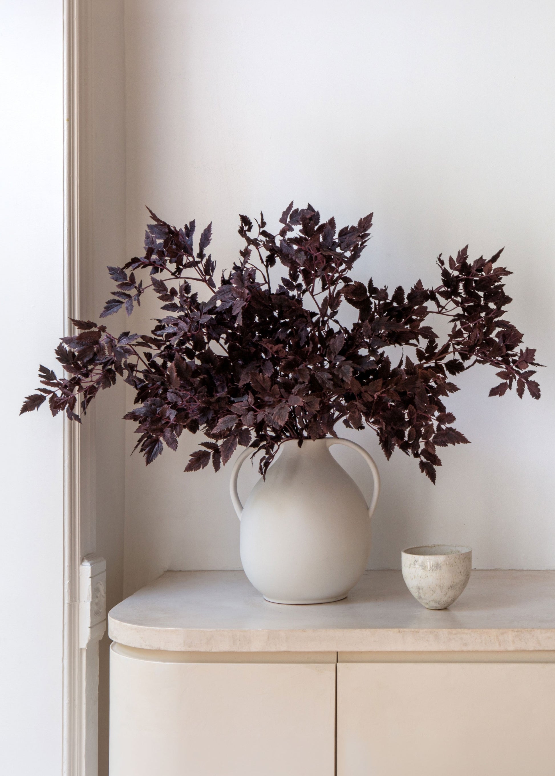 A large white ceramic pitcher vase filled with dark burgundy artificial foliage, displayed on a light beige console table next to a small textured ceramic cup against a white wall.