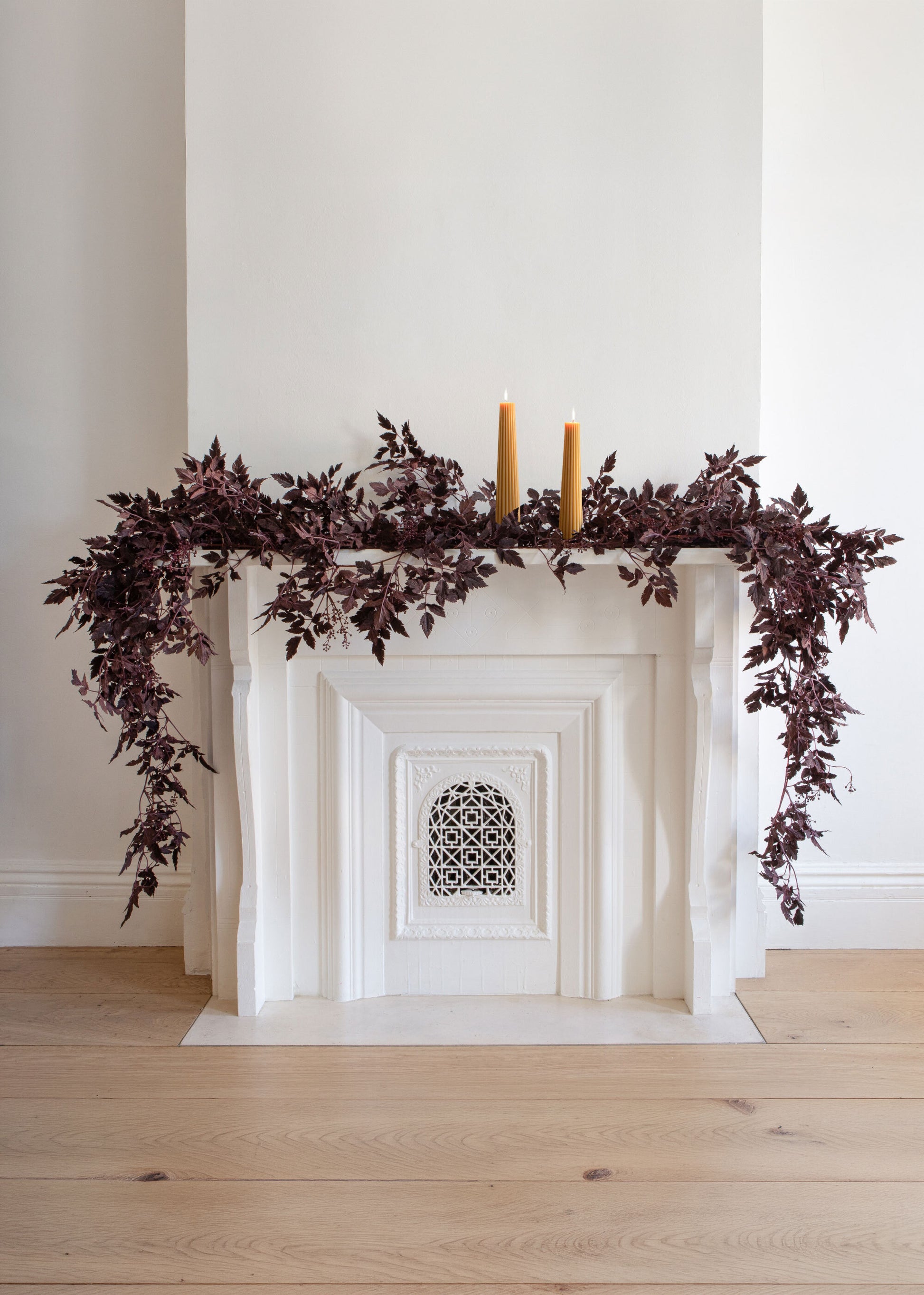 A white fireplace mantel decorated with dark burgundy leafy cimicifuga garland and two lit yellow pillar candles, set against a white wall and light wood floor.