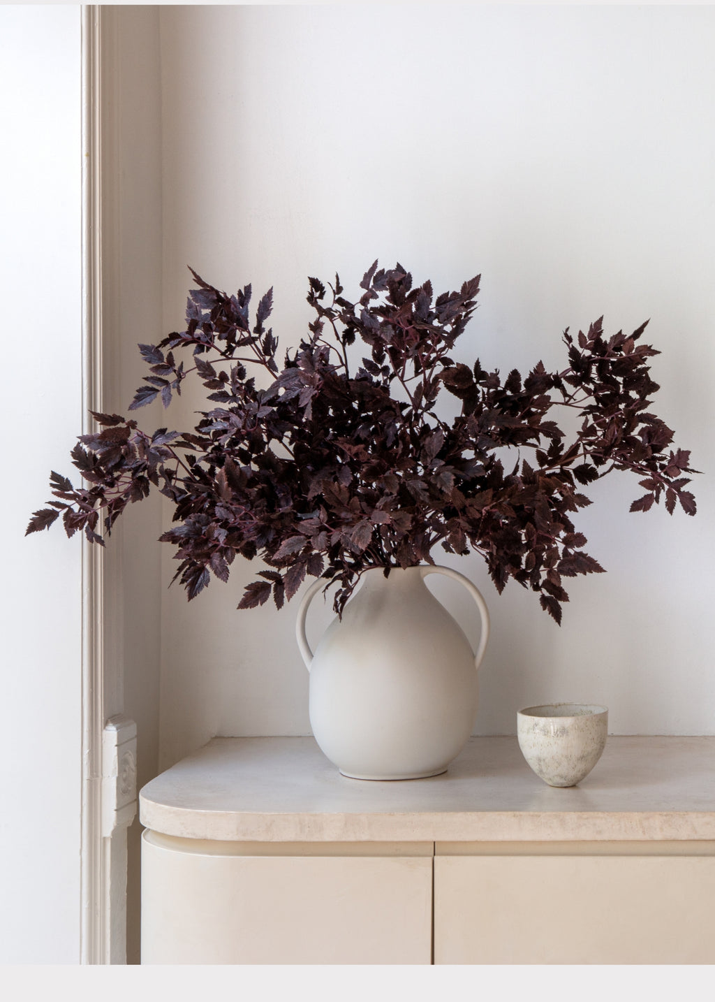 A large white ceramic pitcher vase filled with dark burgundy artificial foliage, displayed on a light beige console table next to a small textured ceramic cup against a white wall.