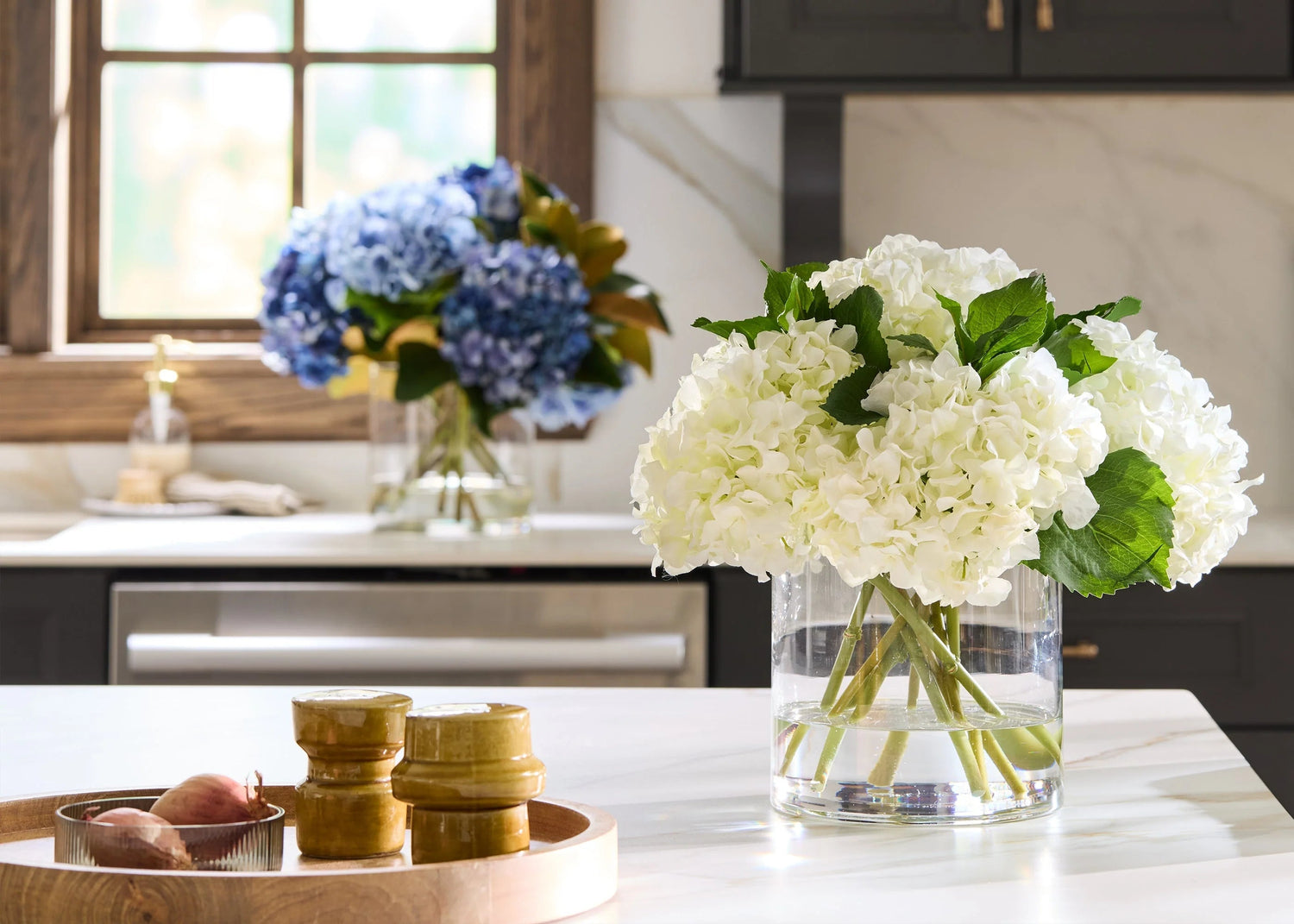 White flowers in a vase on a kitchen counter with a window and cabinets in the background. | Afloral