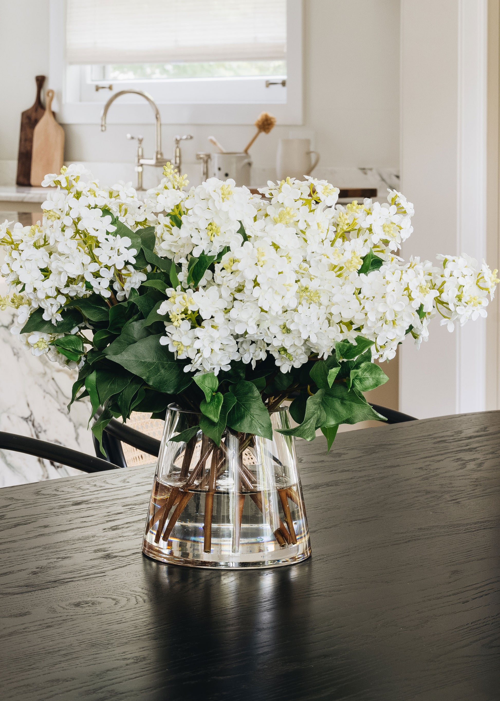 Clear vase with white lilac flowers on a wooden table in a kitchen.