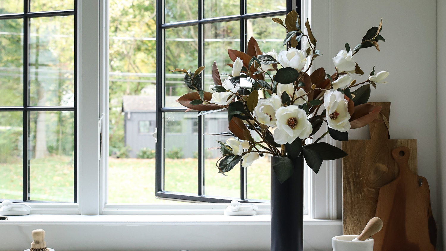 Decorative flower arrangement in a vase on a windowsill with a view of greenery outside.