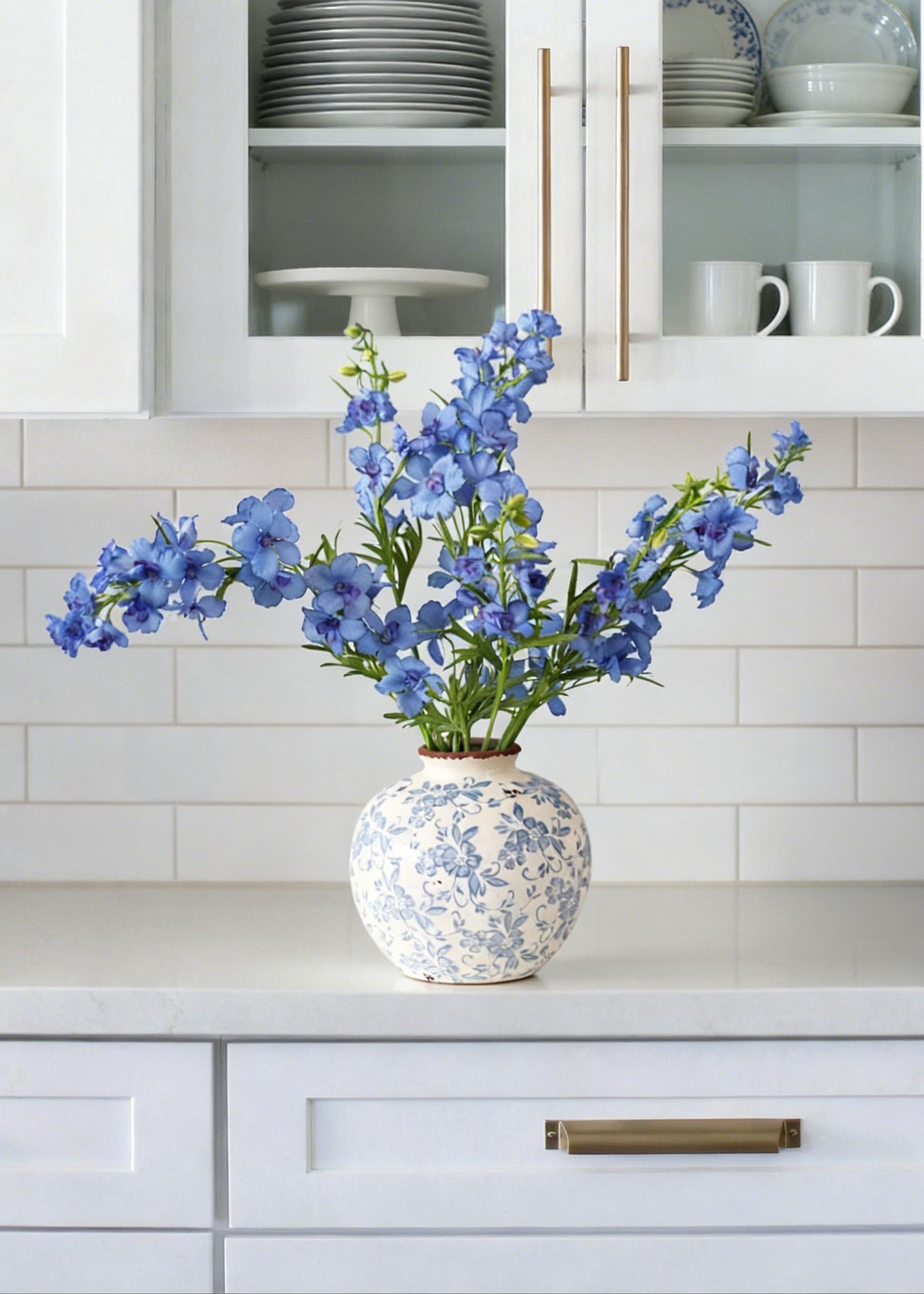 Blue floral arrangement in a decorative vase on a kitchen counter with white cabinets and subway tiles.