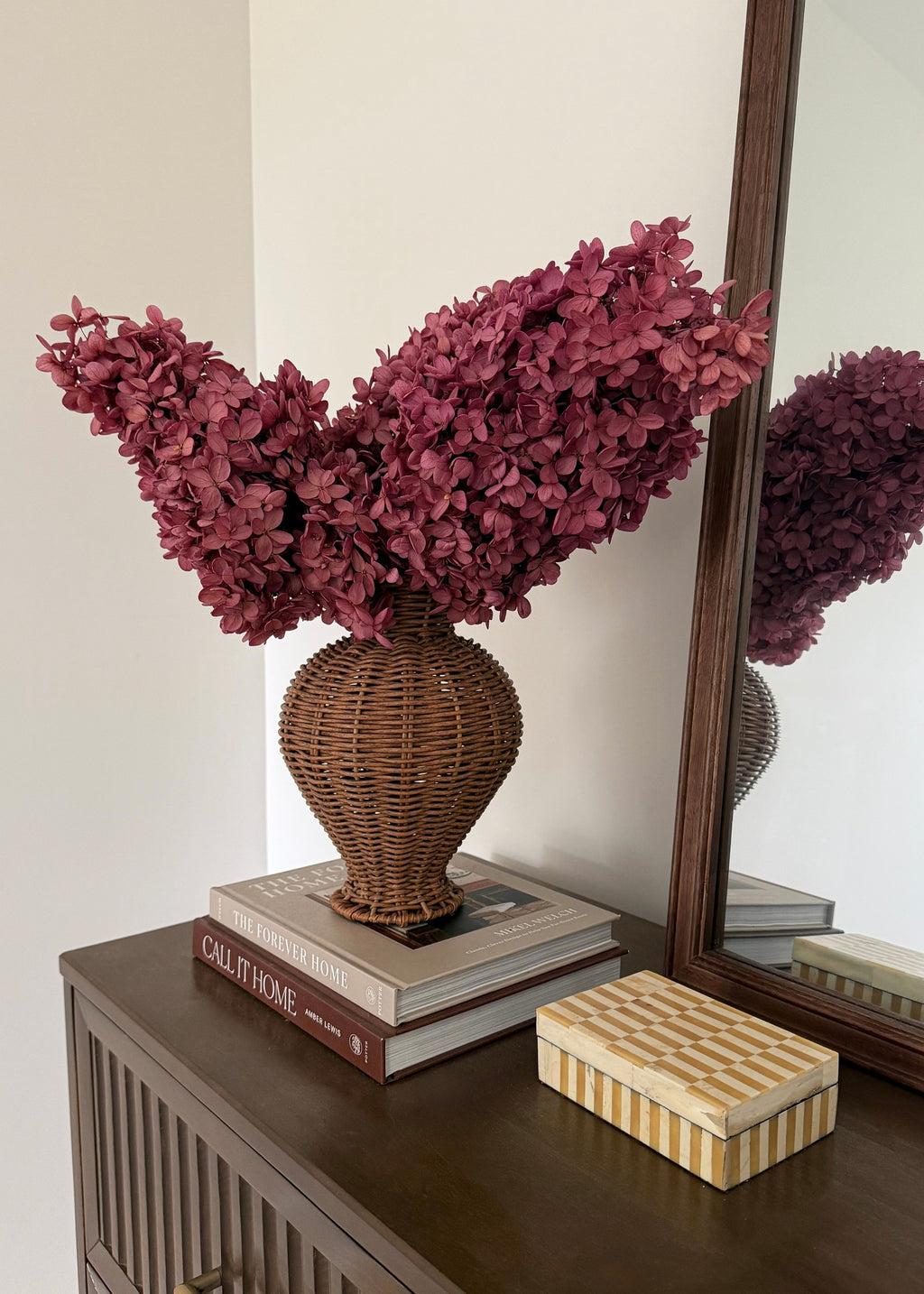 A decorative arrangement of deep burgundy dried hydrangeas in a brown woven wicker vase, displayed on a dark wood console table next to a mirror and a striped decorative box.