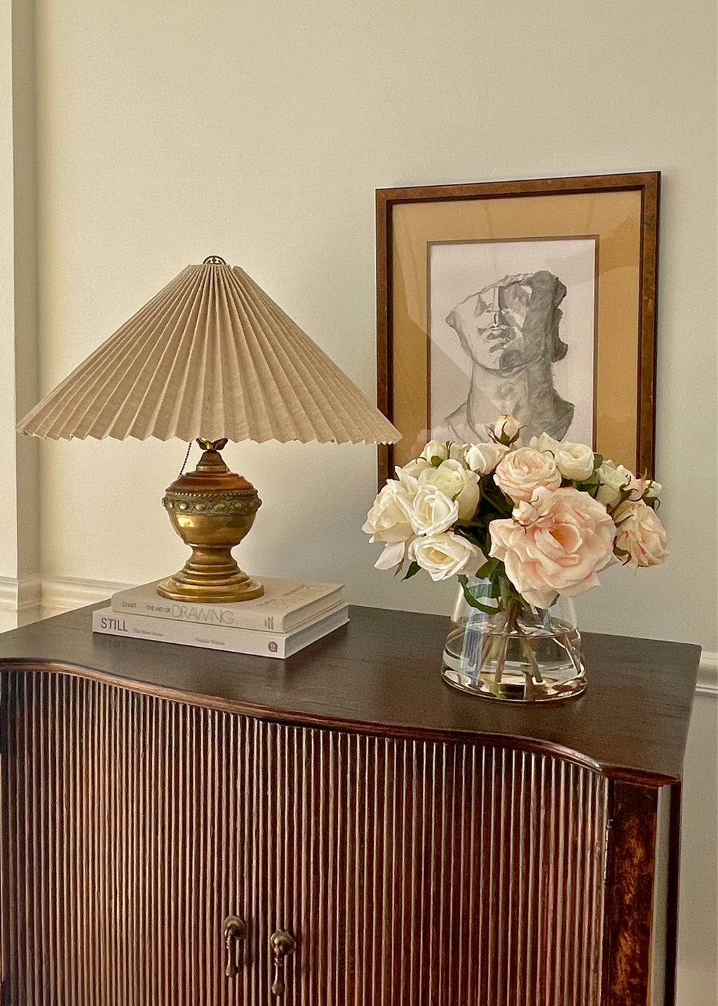 Decorative lamp, vase with flowers, and framed artwork on a wooden cabinet.