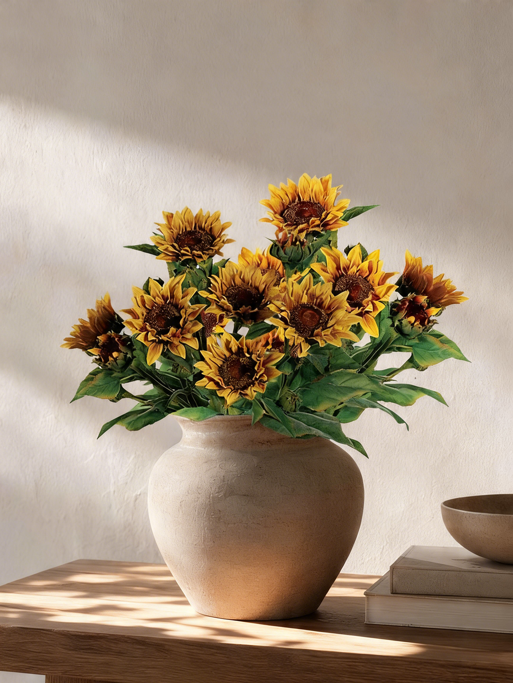 A bouquet of artificial sunflowers with yellow petals and dark brown centers, arranged in a round beige ceramic vase on a wooden table against a textured beige wall.